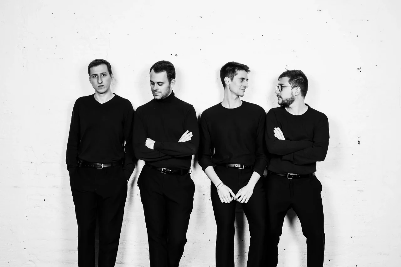 Schwarz-Weiß-Porträt der vier Musiker des Kebyart Saxophonquartetts in dunklen Rollkragenpullovern vor einer hellen Ziegelwand.